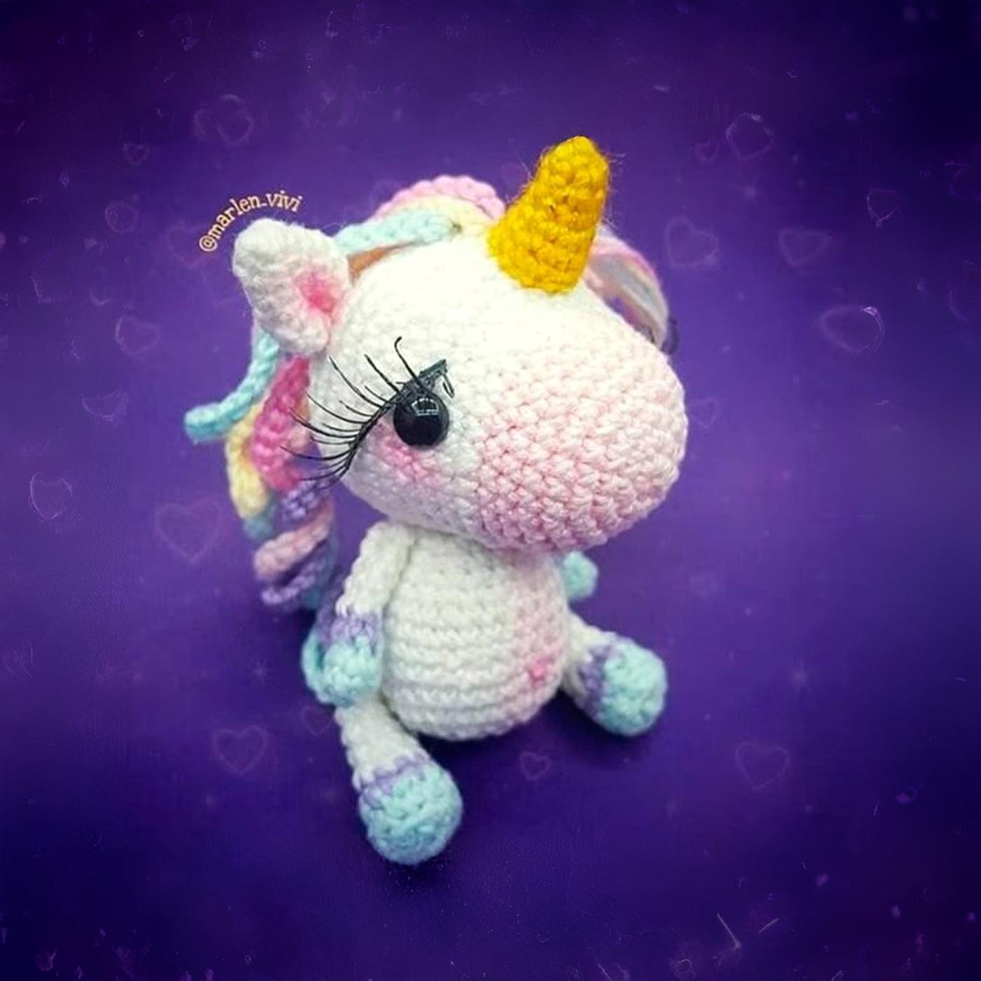 Unicornio amigurumi tejido a ganchillo con cuerpo blanco, crin y cola de colores pastel, y un cuerno amarillo, sentado sobre un fondo claro.