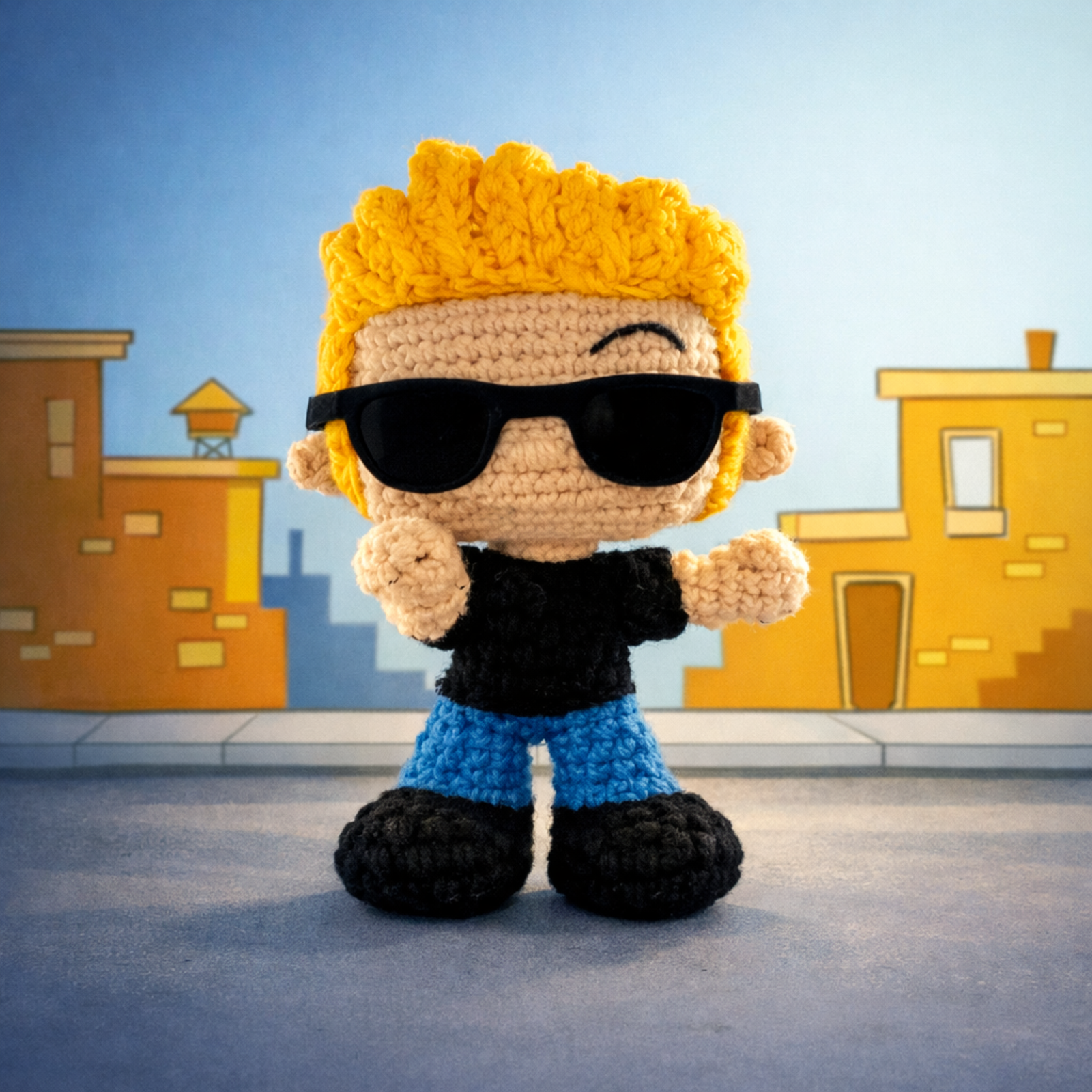 Muñeco amigurumi de un chico con pelo amarillo, gafas de sol negras y vaqueros, posando con confianza en una calle de ciudad.