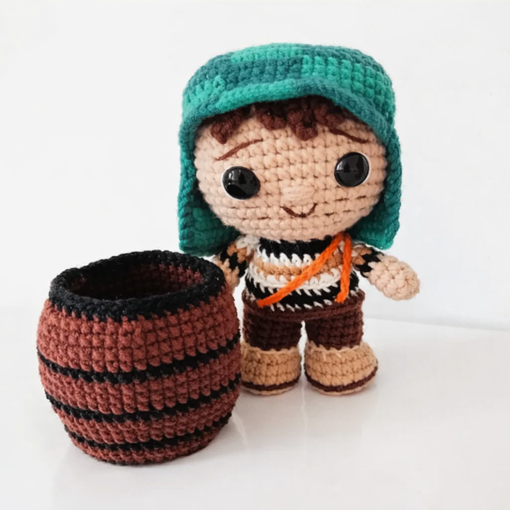 Muñeco amigurumi del Chavo del Ocho con gorro verde y tirantes naranjas, de pie junto a un barril tejido marrón.