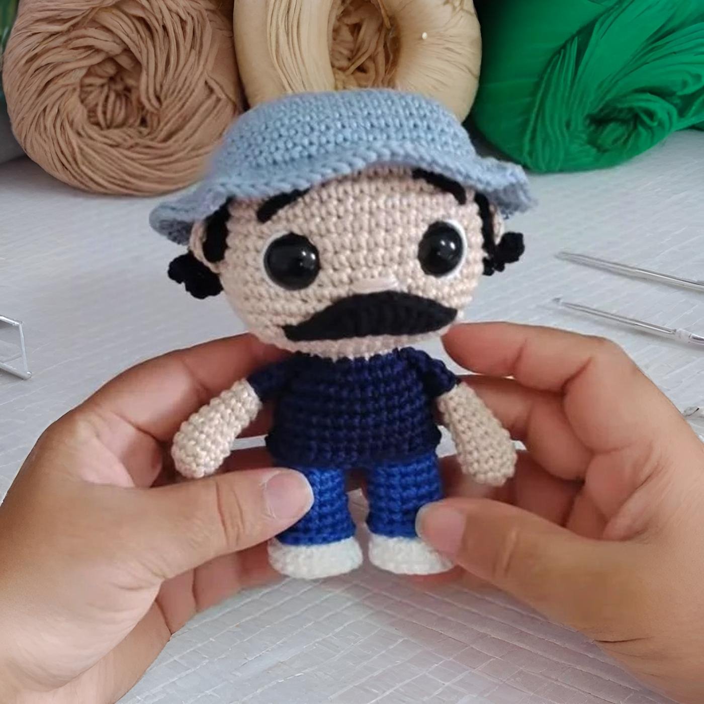 Manos sosteniendo un amigurumi de Don Ramón con su característico gorro celeste, bigote negro y camiseta azul oscura.