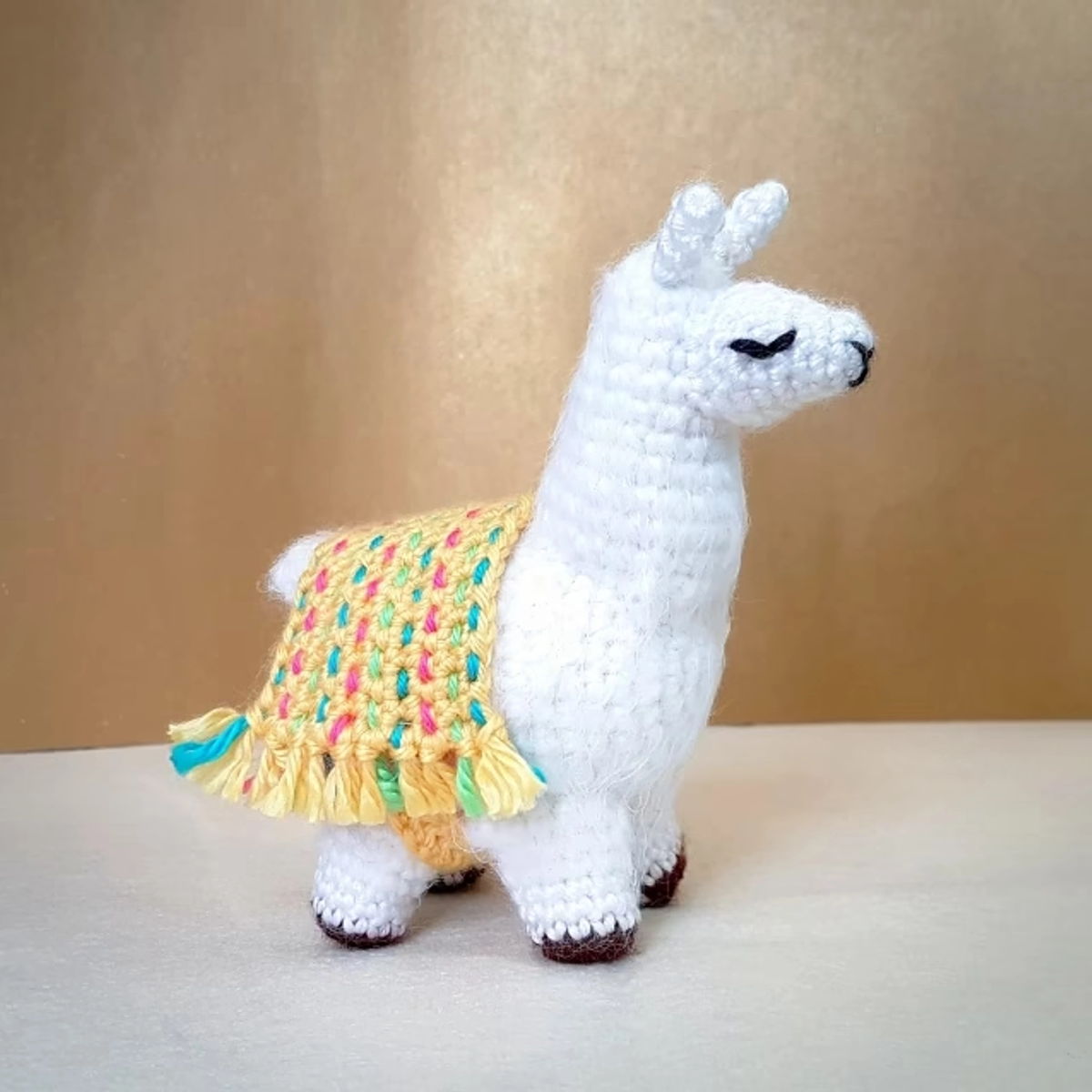 Una llama blanca de ganchillo, de pie y de perfil, con una manta amarilla y colorida sobre su lomo.