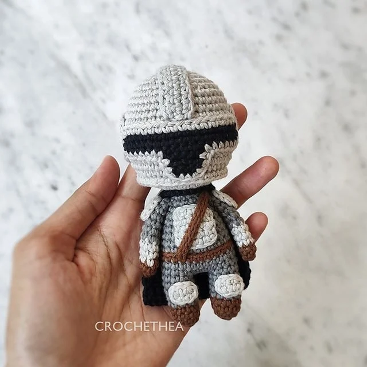 Mano sosteniendo un pequeño muñeco amigurumi del Mandaloriano con su casco y armadura gris tejidos a ganchillo.