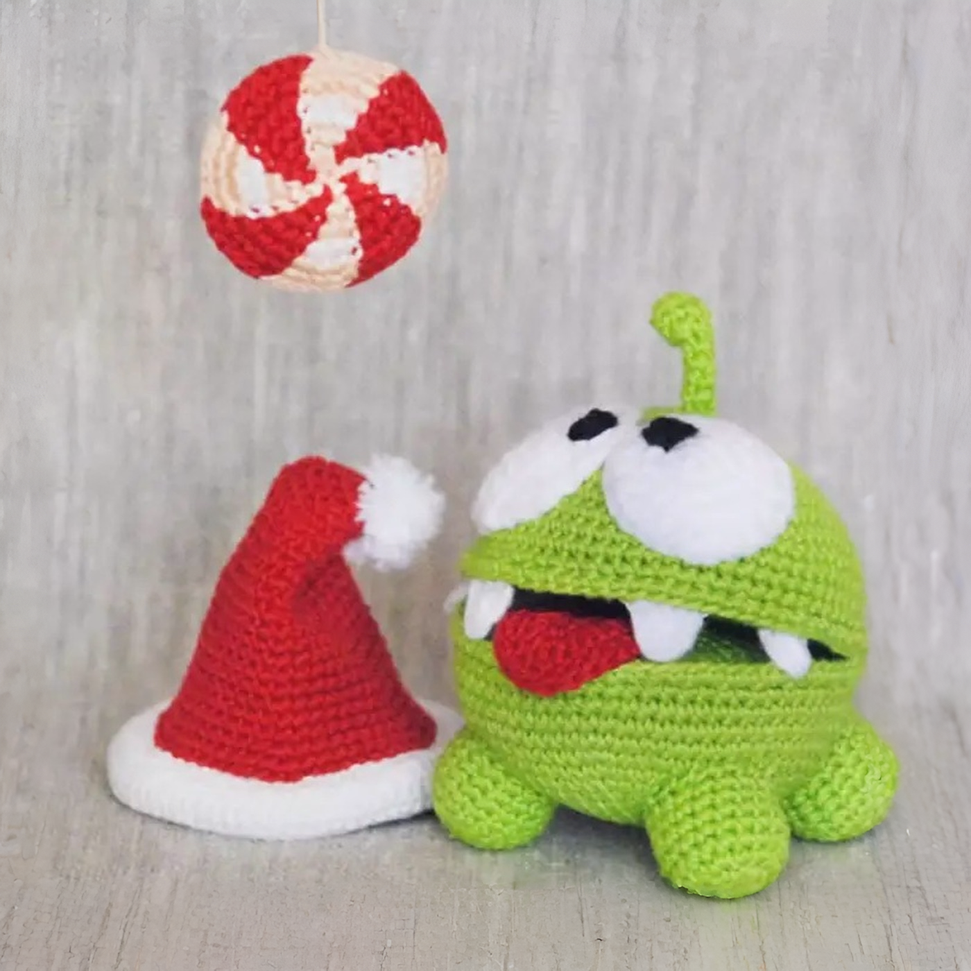Muñeco de ganchillo de Om Nom verde junto a un gorro de Papá Noel y un caramelo colgante a crochet