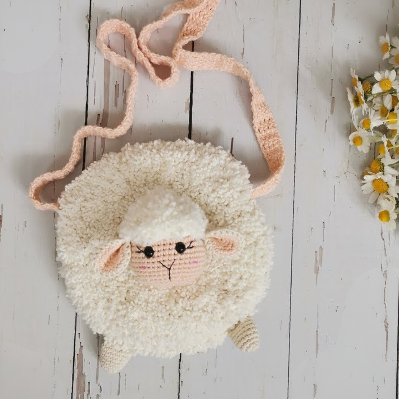 Bolso redondo tejido a crochet con forma de oveja blanca, detalles en rosa y asa de lana sobre fondo de madera blanca.