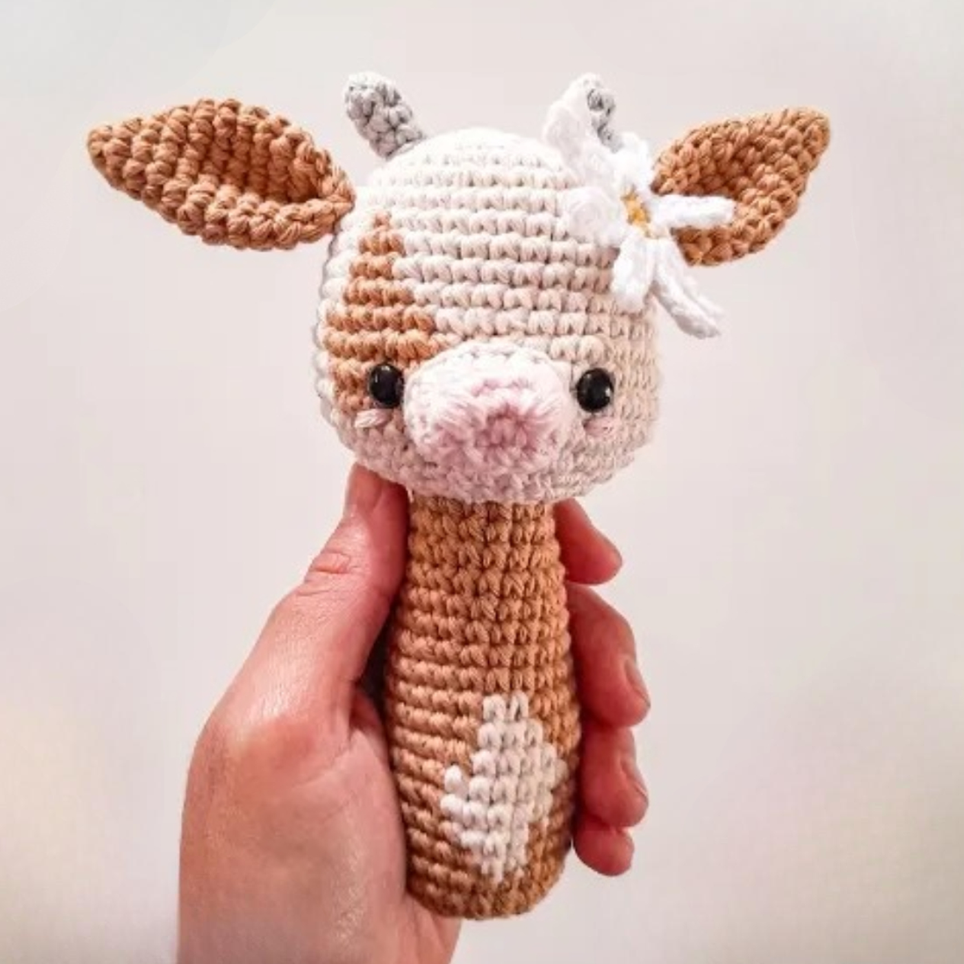 Mano sosteniendo un sonajero de amigurumi con forma de vaca, color beige y blanco con una flor en la oreja.