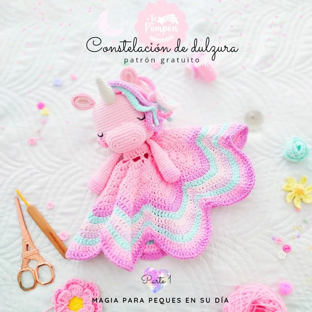 Muñeco amigurumi de unicornio con mantita de crochet en tonos pastel, posado sobre una superficie blanca acolchada.