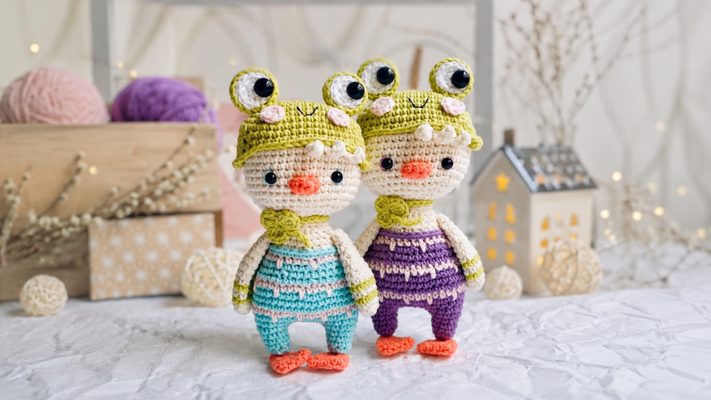 Dos pollitos amigurumi tejidos a crochet, ambos con gorritos verdes de rana, uno con overol azul y otro morado.
