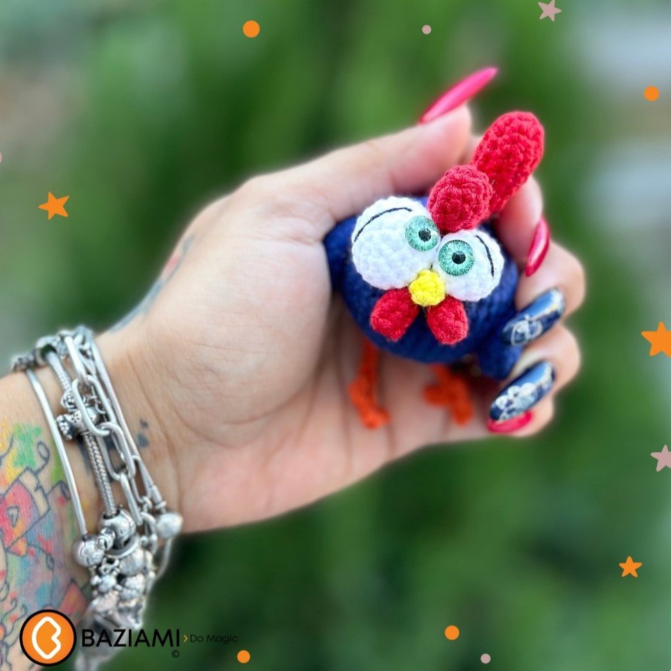 Gallinita amigurumi azul con cresta roja sostenida por una mano sobre un fondo verde desenfocado.