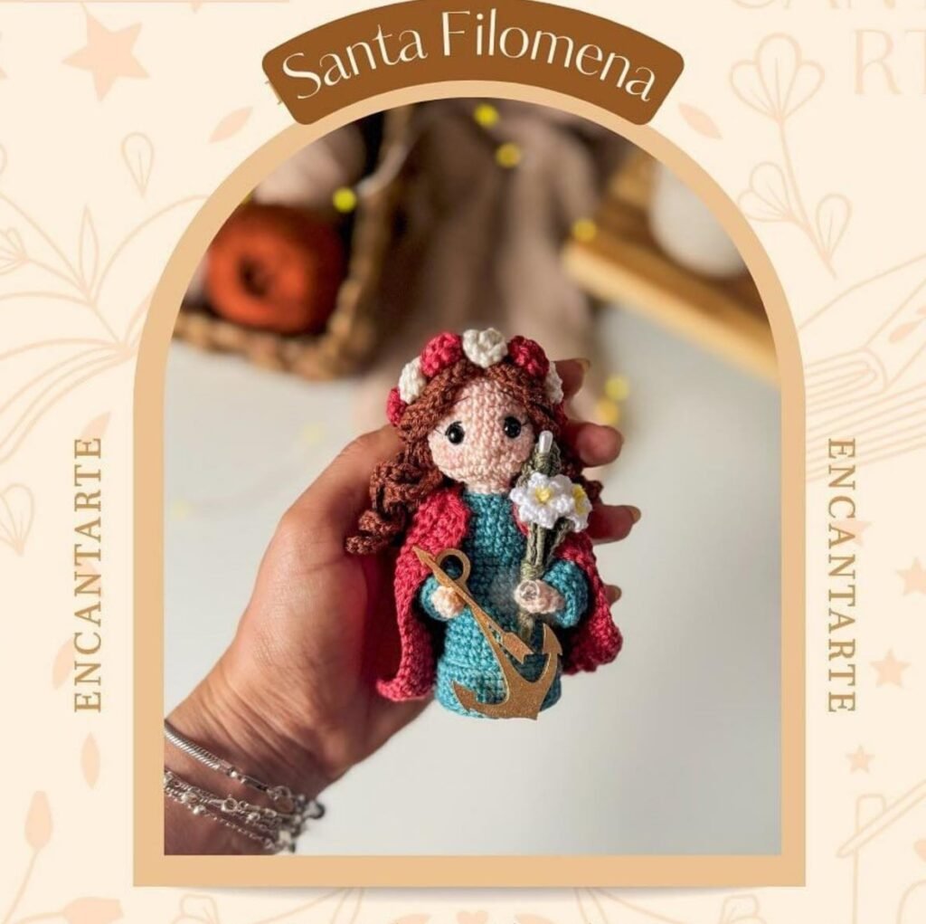 Muñeca amigurumi de Santa Filomena con capa roja, vestido turquesa y una pequeña ancla dorada.