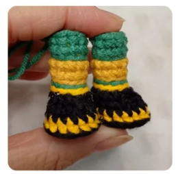Mano sosteniendo un par de botas tejidas a crochet en colores verde, amarillo y negro para el amigurumi Zayu.