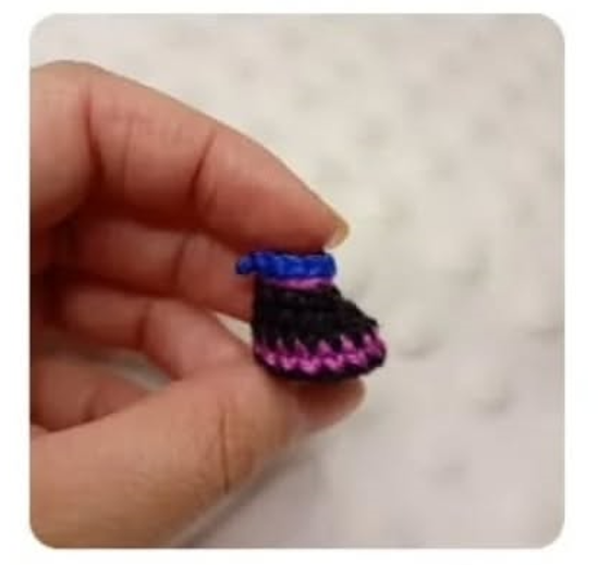 Mano sosteniendo una pequeña pieza de amigurumi en forma de botín tejida con hilo negro, azul y violeta.