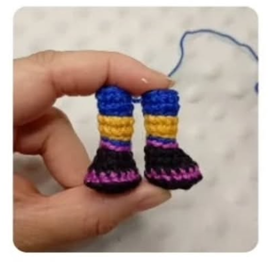 Mano sosteniendo un par de botas tejidas a ganchillo con franjas de colores azul, amarillo, morado y negro.