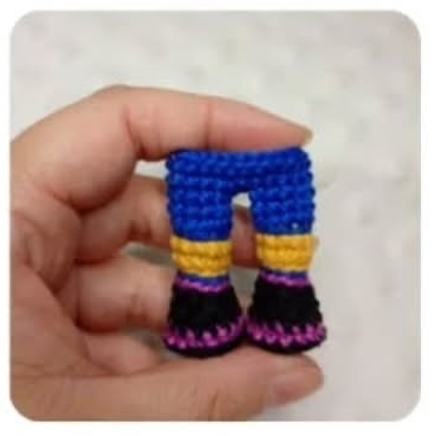 Mano sosteniendo unas pequeñas piernas de amigurumi tejidas en azul, amarillo y negro sobre un fondo claro.