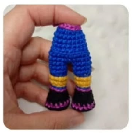 Mano sosteniendo unas pequeñas piernas de amigurumi tejidas en colores azul, amarillo, negro y detalles en fucsia.