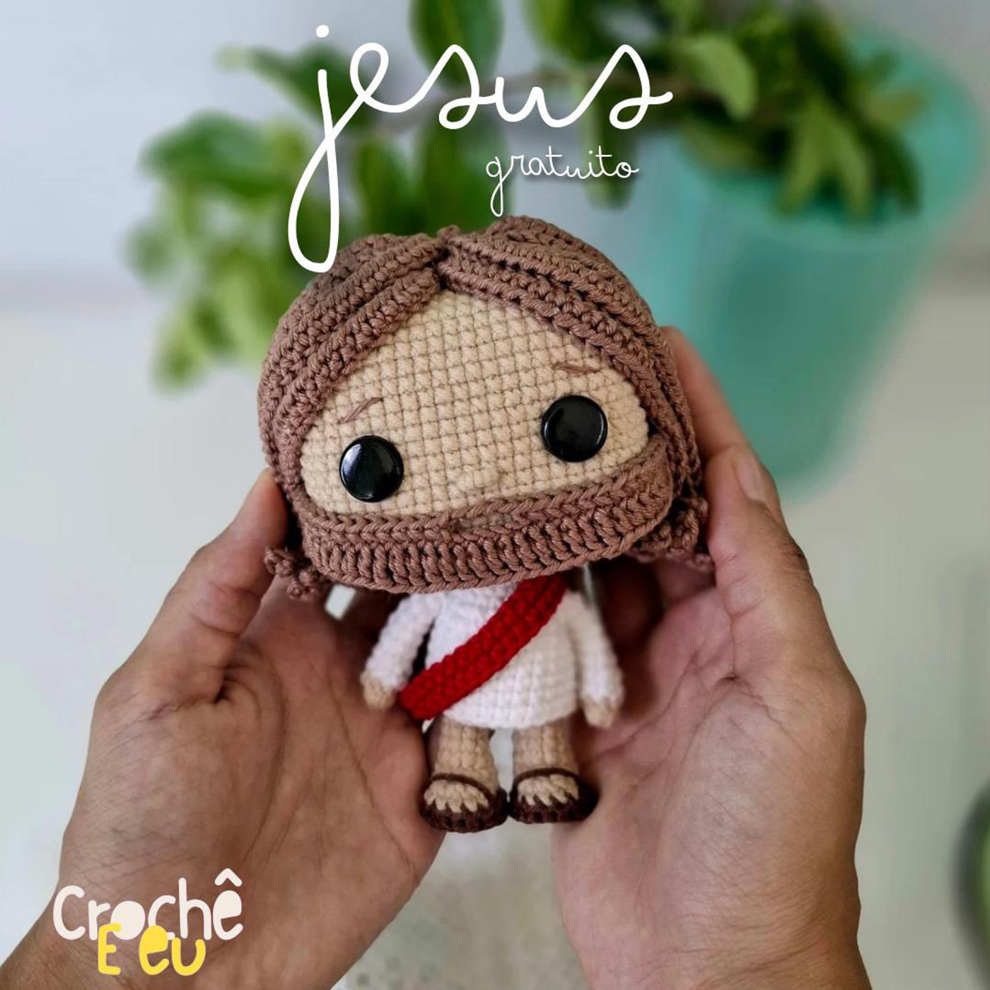 Manos sosteniendo un muñeco amigurumi de Jesús con túnica blanca, banda roja, pelo castaño y ojos de seguridad negros.