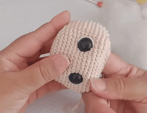 Primer plano de unas manos uniendo los ojos a una cabeza de amigurumi de Don Ramón tejida a crochet.