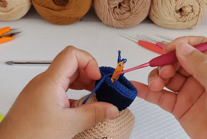 Primer plano de manos tejiendo un amigurumi de Don Ramón, utilizando un hilo azul y beige.