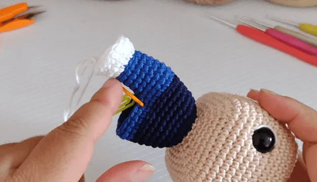 Primer plano de manos tejiendo el cuello de un amigurumi de Don Ramón, mostrando el hilo azul y blanco.