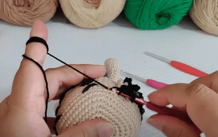 Primer plano de manos tejiendo cabello negro sobre un amigurumi Don Ramón.