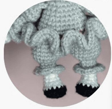 Una Imagen De Primer Plano Muestra Un Objeto Tejido a Crochet Con Detalles De Volantes En Hilo Gris Claro El Objeto Incluye Patas Con Pies Blancos Y Puntas Negras El Fondo Está Suavemente Borroso En Un Tono Rosa Claro