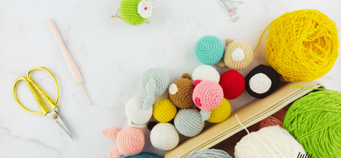 Una variedad de coloridas bolas de crochet y otros pequeños amigurumis se muestran cerca de un ovillo de hilo amarillo, tijeras, un ganchillo y algunos alfileres de costura. El fondo es una superficie blanca y todos los elementos están colocados de forma organizada.