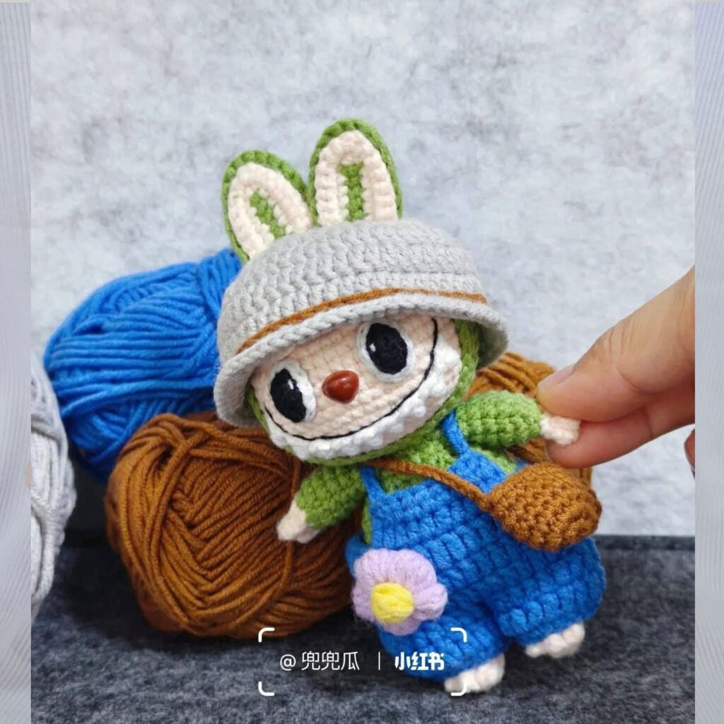Una Mano Sostiene Un Muñeco Labubu Tejido a Crochet Con Orejas De Conejo Que Lleva Un Sombrero Gris Un Mono Azul Y Una Camisa Verde Frente a Bolas De Hilo