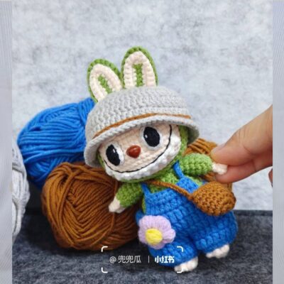 Una Mano Sostiene Un Muñeco Labubu Tejido a Crochet Con Orejas De Conejo Que Lleva Un Sombrero Gris Un Mono Azul Y Una Camisa Verde Frente a Bolas De Hilo