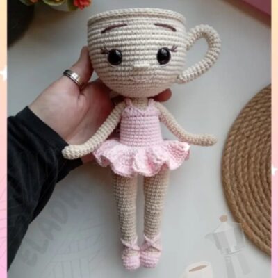 Muñeca Amigurumi Crochet De Ballerina Capuchina Con Cabeza De Taza Beige Ojos Negros Y Sonrisa Viste Tutú Rosa Y Zapatillas De Ballet Sostenida Por Una Mano Con Flores Y Posavasos Tejidos Al Fondo Ideal Para Fans Del Amigurumi Creativo
