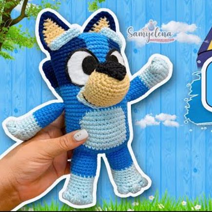Una Mano Sostiene Un Juguete De Peluche De Perro Amigurumi Bluey De Ganchillo Contra Un Fondo De Madera Azul Con Hierba En La Parte Inferior El Perro Tiene Ojos Grandes Nariz Negra Y Brazos Levantados El Fondo Presenta Algunas Hojas Y Ramas Y Un Logo Que Dice samyelena