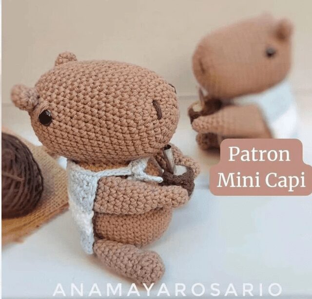 Un Capibara En Miniatura Tejido a Crochet Se Sienta Sosteniendo Un Pequeño Instrumento Mientras Que Otra Figura Similar Se Ve Al Fondo Al Lado Se Muestra El Texto patrona Mini Capi Y En La Parte Inferior De La Imagen Está Escrito anamayarosario