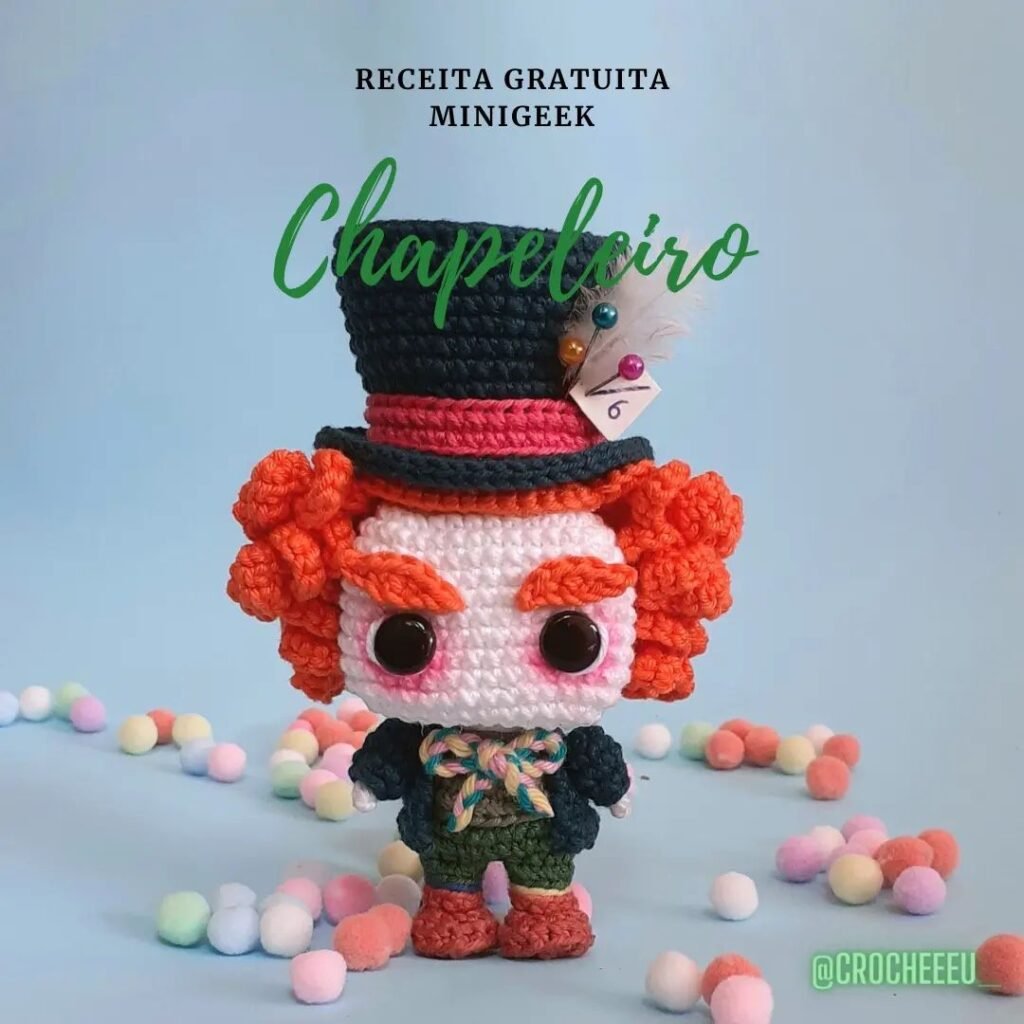 Muñeco amigurumi de un personaje de pelo rizado naranja, ojos grandes y chistera negra adornada con diversos adornos. La muñeca está vestida con un atuendo colorido y caprichoso. A su alrededor hay pequeños pompones de colores pastel. El texto de arriba dice "Receita Gratuita Minigeek Chapeleiro - tutorial amigurumi español.