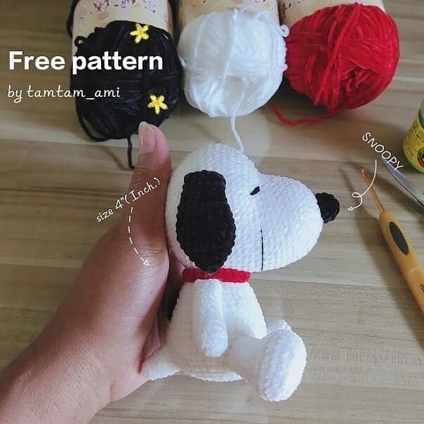 Una mano sostiene un pequeño amigurumi de Snoopy tejido a crochet sobre una superficie de madera. Al fondo hay ovillos de hilo en negro, blanco y rojo, junto con un ganchillo. El texto dice “Patrón de crochet gratis” de tamtam_ami e indica el tamaño de la figura como 4 pulgadas.