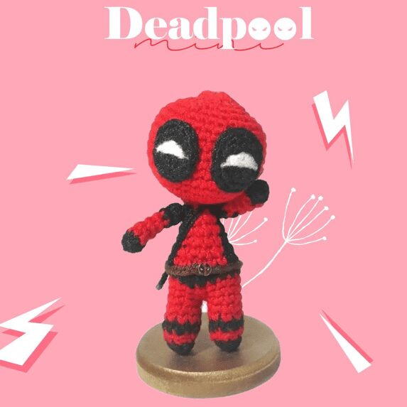 Una Figura Tejida a Crochet De Deadpool Se Alza Sobre Una Base Redonda Sobre Un Fondo Rosa La Figura Que Muestra Un Patrón Amigurumi Deadpool Tiene Un Traje Rojo Y Negro Grandes Ojos Blancos Y Posa Con Un Brazo Doblado La Palabra deadpool Está Escrita En La Parte Superior Rodeada De Relámpagos Blancos Y Gráficos En Forma De Garabatos