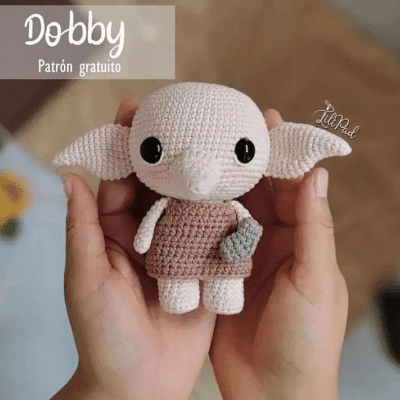 Un par de manos sostiene un muñeco de crochet que se parece a Dobby de Harry Potter. La muñeca, tejida a crochet, presenta grandes orejas caídas, grandes ojos negros y un vestido sencillo. El texto de la imagen dice "Dobby Patrón Amigurumi gratuito" con un logo en la esquina superior derecha.