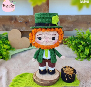 Un muñeco de crochet hecho a mano de un duende de la fortuna con cabello naranja y un traje verde se alza sobre una base redonda de madera. Al lado de la muñeca hay una pequeña olla negra llena de paja. El fondo incluye follaje verde y una decoración de corazón, todo hecho con un patrón gratuito.