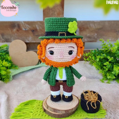 Un muñeco de crochet hecho a mano de un duende de la fortuna con cabello naranja y un traje verde se alza sobre una base redonda de madera. Al lado de la muñeca hay una pequeña olla negra llena de paja. El fondo incluye follaje verde y una decoración de corazón, todo hecho con un patrón gratuito.