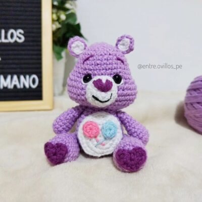 Un oso morado de ganchillo con una cara sonriente se sienta sobre una superficie suave. Tiene color blanco dentro de las orejas y en el vientre, el cual está adornado con un corazón azul y rosa. Un ovillo de hilo morado está a la derecha y un cartel parcialmente visible que ofrece patrón gratuito para tejido amigurumi está a la izquierda.