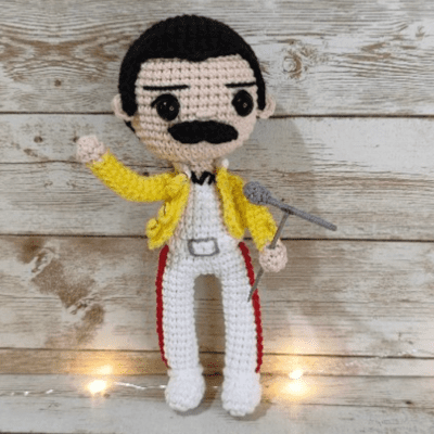 Un muñeco de crochet que se parece a Freddie Mercury, con bigote, cabello negro y ojos expresivos, que viste una chaqueta de color amarillo brillante sobre un traje blanco con detalles en rojo y negro y sostiene un micrófono gris. El fondo son tablones de madera rústicos con luces amarillas en el suelo.