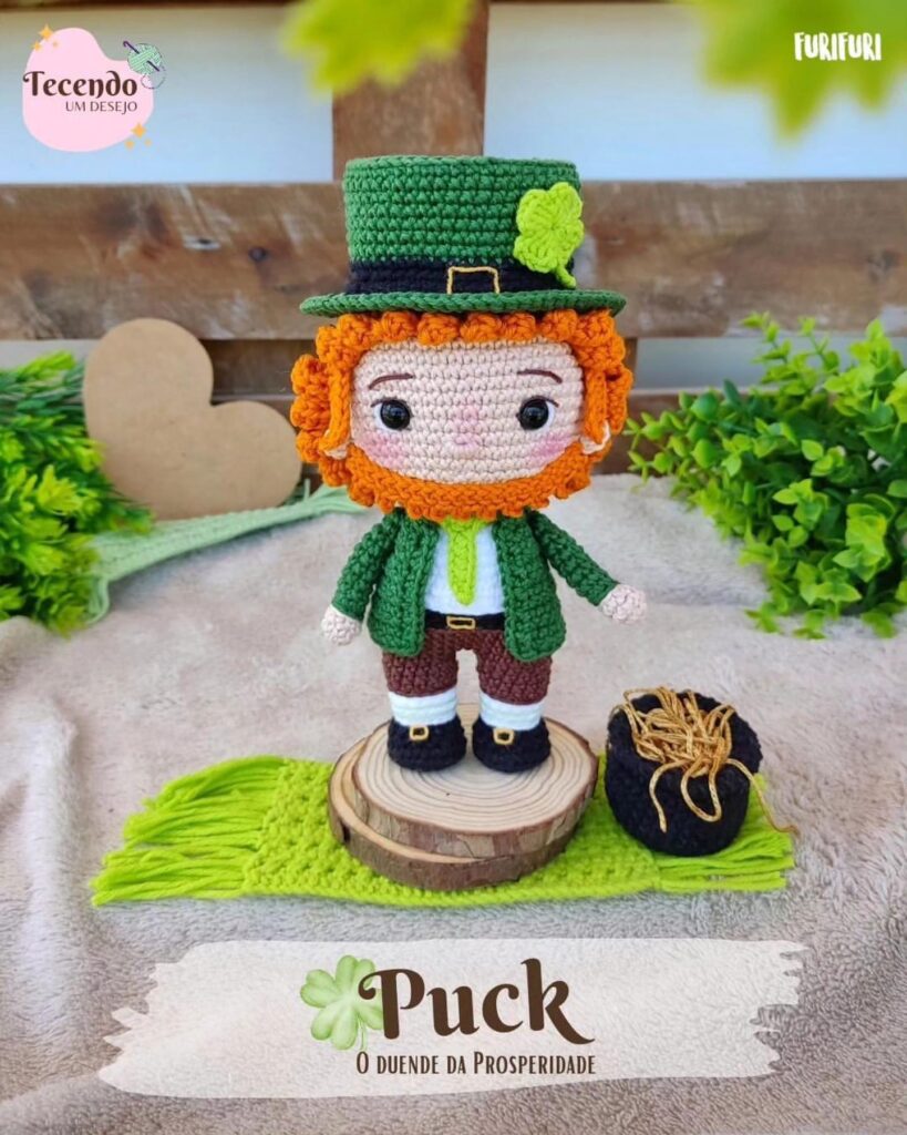En Un Disco De Madera Se Muestra Un Muñeco Amigurumi Tejido a Crochet Con Una Barba De Color Naranja Brillante Y Un Traje Verde Que Se Asemeja a Un Duende La Figura Se Encuentra Sobre Una Estera De Punto Verde Junto a Un Saco De Hilo El Texto De La Imagen Dice puck El Duende De La Fortuna El Fondo Incluye Vegetación Y Un Panel De Madera