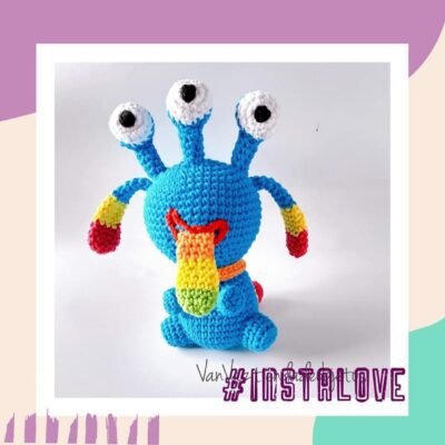 Un monstruo azul tejido a crochet con tres ojos en tallos, un hocico amarillo y rojo y brazos y piernas azules con puntas rojas. Este monstruo colorido amigurumi se asienta sobre una superficie blanca dentro de un marco blanco. El texto en la esquina inferior derecha dice "#INSTALOVE" en violeta.