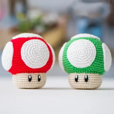 Dos figuras de crochet que parecen setas de una popular serie de videojuegos. Uno tiene una gorra roja con manchas blancas y el otro tiene una gorra verde con manchas blancas. Ambos tienen tallos de color beige con ojos negros y bocas pequeñas. Fondo borroso con varios objetos.