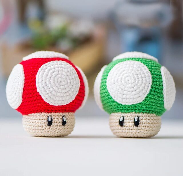 Dos Figuras De Crochet Que Parecen Setas De Una Popular Serie De Videojuegos Uno Tiene Una Gorra Roja Con Manchas Blancas Y El Otro Tiene Una Gorra Verde Con Manchas Blancas Ambos Tienen Tallos De Color Beige Con Ojos Negros Y Bocas Pequeñas Fondo Borroso Con Varios Objetos