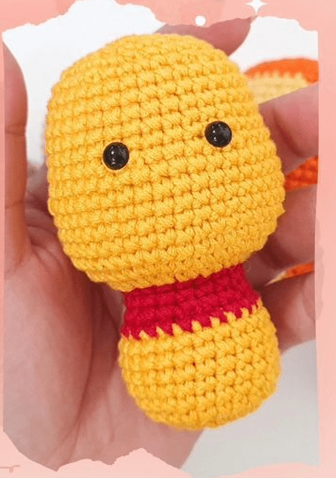 Una Mano Sostiene Una Pequeña Figura De Amigurumi Amarilla Con Cuello Rojo Y Dos Ojos Negros La Figura Que Se Parece a Winnie Pooh Tiene Una Cabeza De Forma Ovalada Y Un Cuerpo Redondo