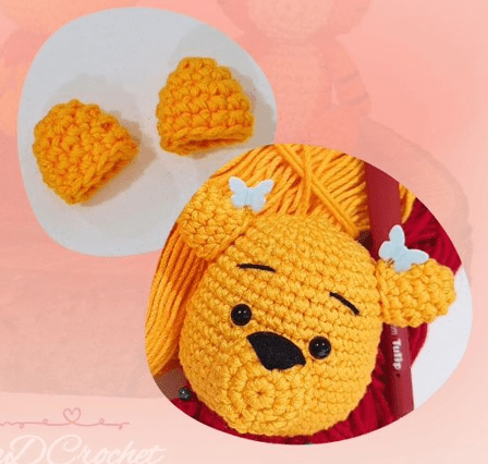 Una Cabeza De Oso Tejida a Crochet Con Hilo Amarillo Que Muestra Ojos Y Nariz Bordados En Negro Y Dos Pequeños Lazos Blancos En Las Orejas Sobre La Cabeza Del Oso Se Encuentran Dos Pequeños Conos Tejidos En Amarillo Al Fondo Se Pueden Ver Herramientas De Crochet E Hilo perfecto Para Un Patrón Amigurumi De Winnie Pooh