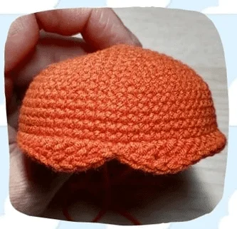 Una Mano Sostiene Una Pieza De Crochet Naranja Con Forma Redonda Y Ligeramente Abovedada La Pieza Tiene Un Patrón Texturizado Con Un Borde Festoneado En La Parte Inferior