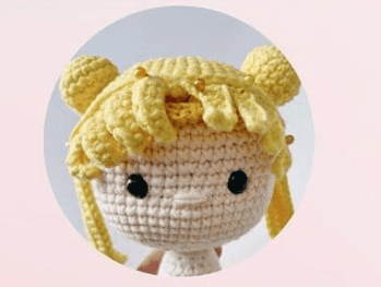 Una Muñeca De Crochet Con Cabello Amarillo Peinado En Dos Moños Y Flequillo Parecido a Sailor Moon Tiene Grandes Ojos Negros Y Una Expresión Simple El Fondo Es De Color Rosa Claro Lo Que Proporciona Un Contraste Encantador Esta Creación De Tejido Bellamente Elaborada Viene Con Un Patrón Gratuito Para Que Lo Pruebes En Casa