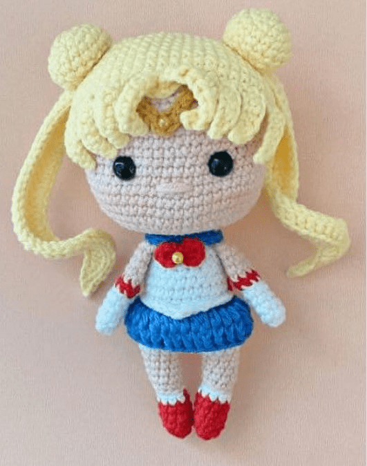 Una Muñeca Amigurumi Sailor Moon Tejida a Crochet Que Se Parece Al Querido Personaje De Anime Viste Una Falda Azul Una Blusa Blanca Con Un Lazo Rojo Y Tiene El Pelo Largo Y Amarillo Peinado En Coletas La Muñeca Tiene Grandes Ojos Negros Y Zapatos Rojos Lo Que Muestra Una Detallada Artesanía En Hilo Y Un Intrincado Tejido