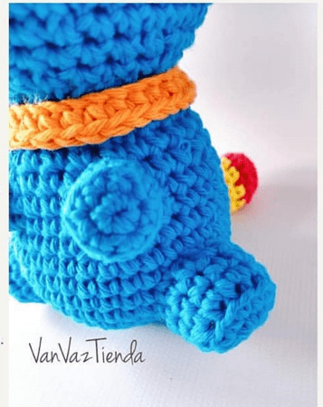 Primer Plano De Una Figura De Juguete Amigurumi Azul Con Un Collar Naranja Visible Y Un Pequeño Apéndice En Su Costado Parte De La Pierna Y Una Sección Multicolor En Forma De Bola Se Suman Al Ambiente De Encanto Monstruo Colorido El Fondo Es Blanco Y La Marca De Agua vanvaztienda Está Escrita En La Parte Inferior Izquierda