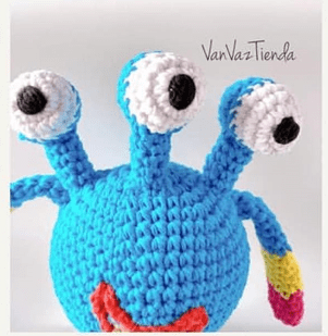 Un Extraterrestre Amigurumi De Crochet Azul Hecho a Mano Con Tres Ojos Altos En Forma De Tallo Y Una Amplia Sonrisa Roja El Cuerpo Redondo Del Extraterrestre Es Suave Con Los Ojos En Diferentes ángulos La Criatura Presenta Brazos Multicolores Azules Y Amarillos vanvaztienda Aparece Al Fondo Junto a Un Patrón Gratuito Para Esta Creación De Tejer