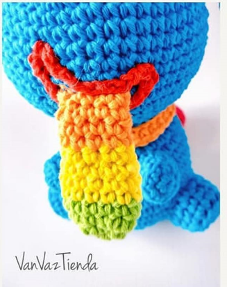 Un Peluche Amigurumi Tejido a Crochet Con Hilo Azul Presenta Una Prominente Cola Multicolor En Tonos De Rojo Naranja Amarillo Verde Claro Y Verde Oscuro Fotografiado Desde Arriba Luce Una Etiqueta Con El Texto vanvaztienda Perfecto Para Quienes Aman Tejer Y Siguen Un Patrón Gratuito